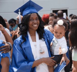 Photo of student in cap and gown carrying small child. 