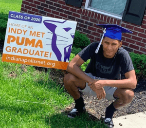 Photo of student in graduation cap by Indy Met yard sign. 